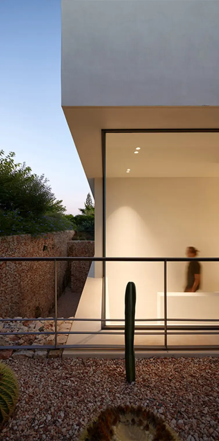 View of a minimalist concrete façade with floor-to-ceiling glass and a cactus garden at dusk in Menorca.
