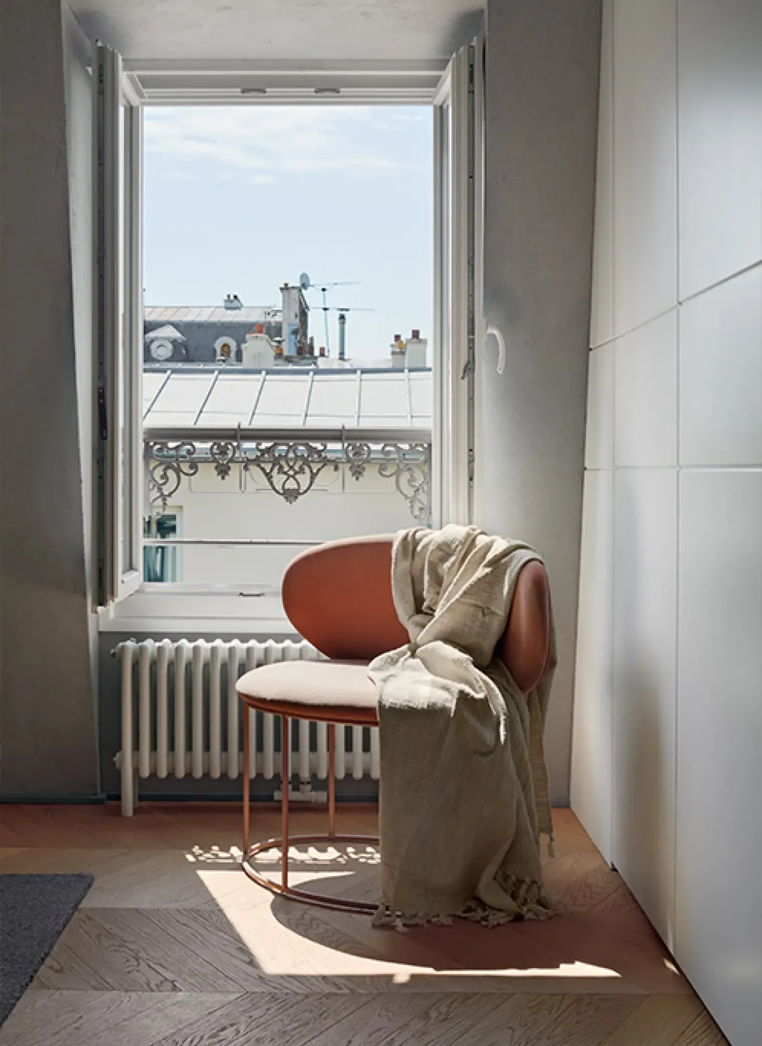Cozy reading corner with terracotta armchair and linen throw beside a window overlooking Parisian rooftops and ornate balcony railings.