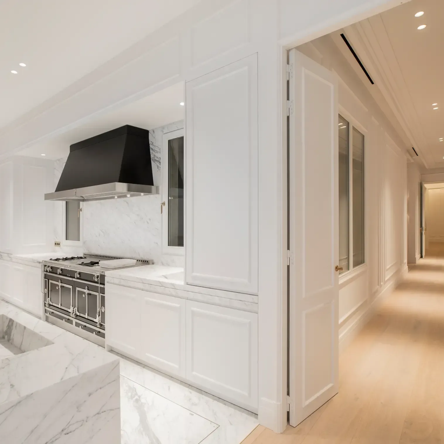 Bright kitchen with marble countertops, a large black hood, and white cabinetry connecting to a warm wood-floor hallway.