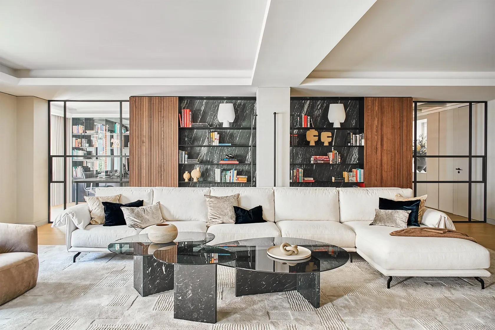 Modern living room with cream sofa and glass coffee tables, flanked by open bookshelves with marble backing and sculptural decor by LUV Studio.