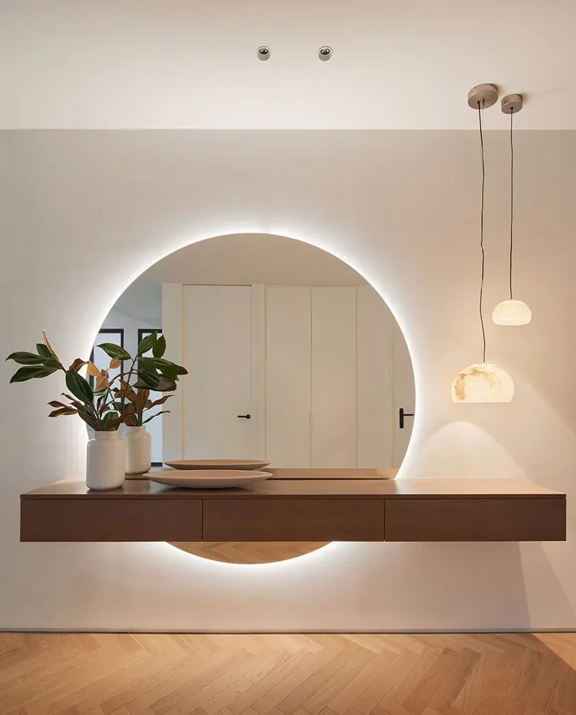 Floating wooden console with round backlit mirror and pendant lights in a luxury apartment entrance by LUV Studio.