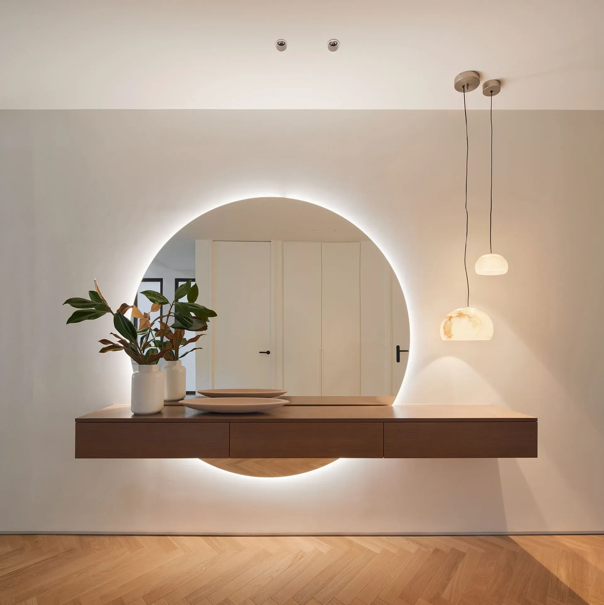 Floating wooden console with round backlit mirror and pendant lights in a luxury apartment entrance by LUV Studio.