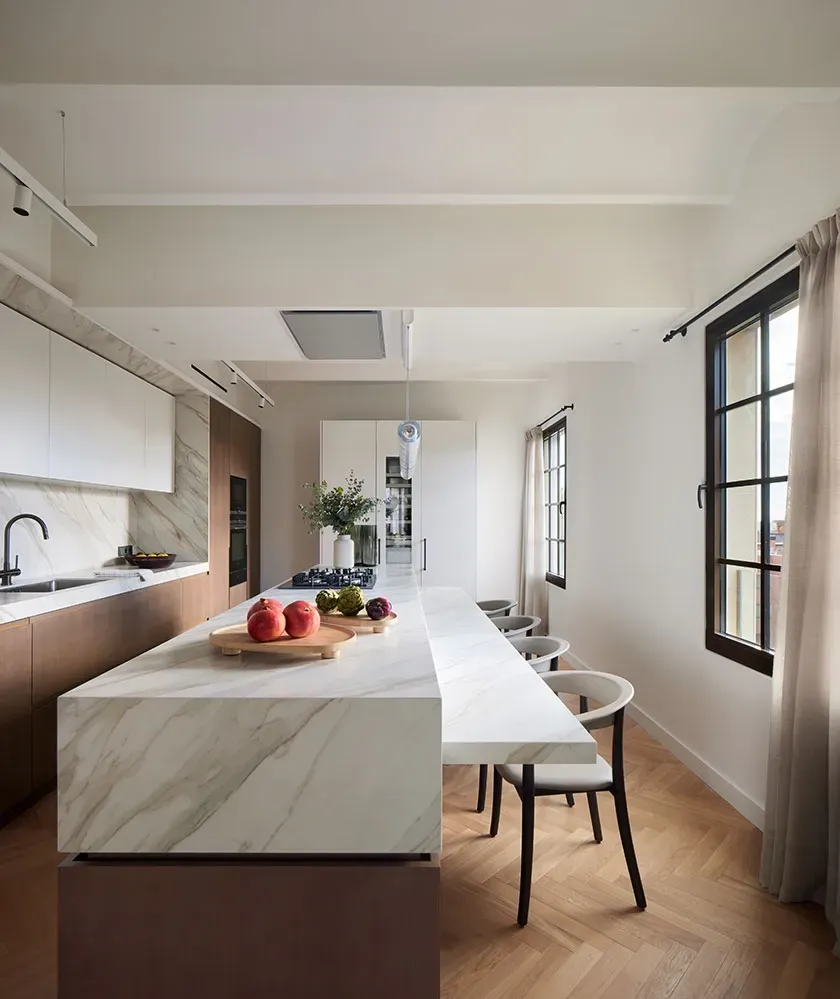 Long marble island with seating and built-in cooktop in a bright, luxury kitchen with natural light.