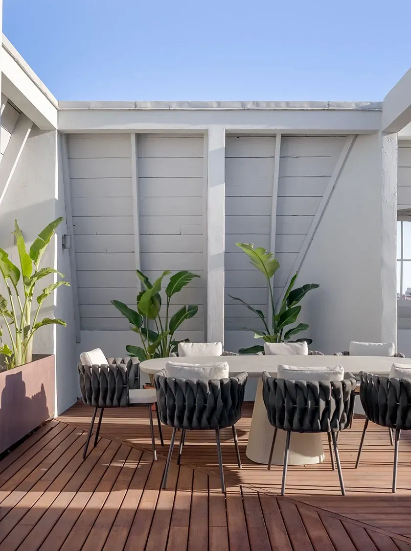 Sunny rooftop terrace with wooden deck, white dining table and woven chairs in a luxury outdoor setting.