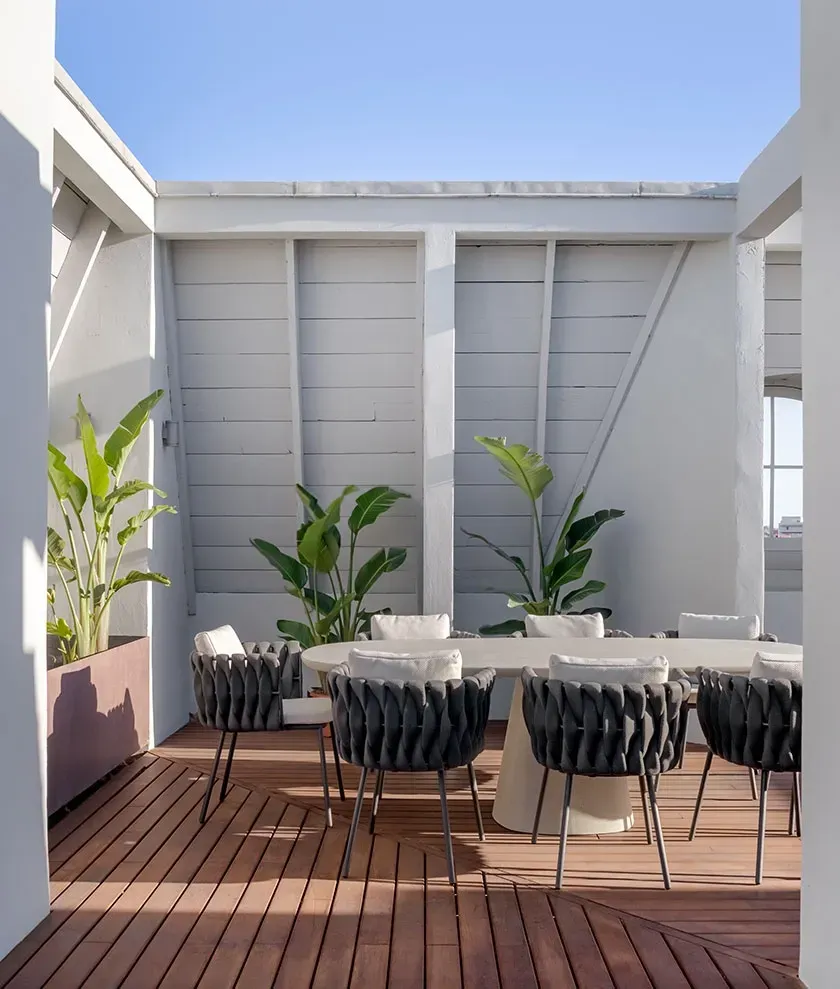 Sunny rooftop terrace with wooden deck, white dining table and woven chairs in a luxury outdoor setting.