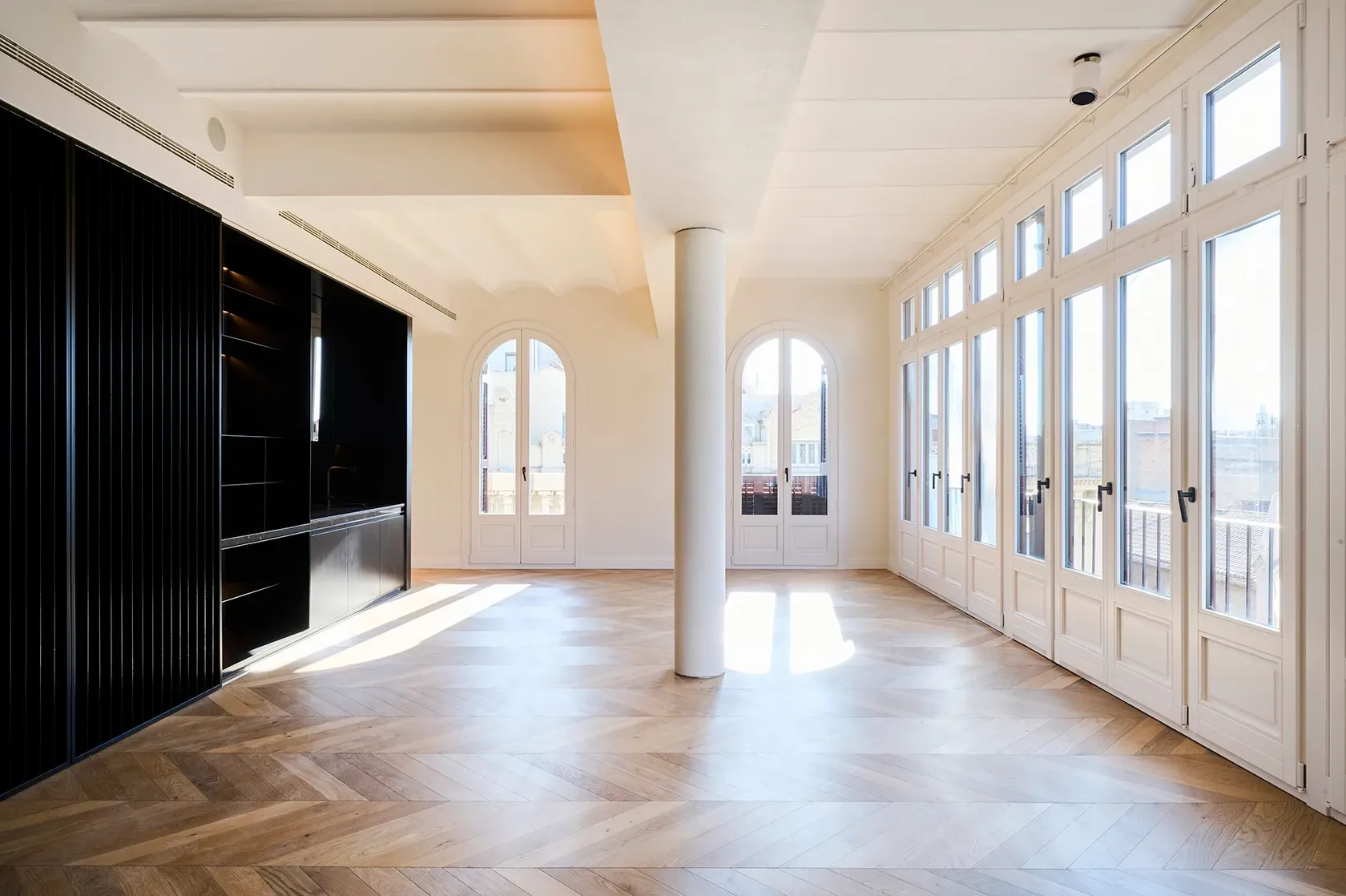 Panoramic view of a bright interior with arched windows, black shelving, and a central white column, blending historic charm and modernity.