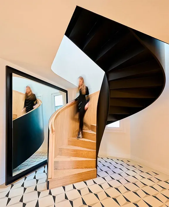 Woman descending a sculptural spiral staircase with oak steps and black steel railing, surrounded by bold geometric floor tiles and natural light.