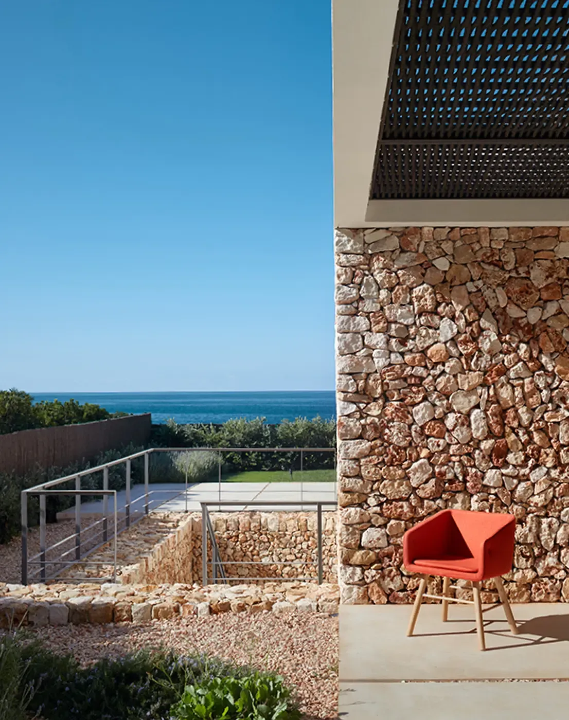Exterior view of a Mediterranean stone wall with a red chair and ocean backdrop, blending coastal and contemporary design.