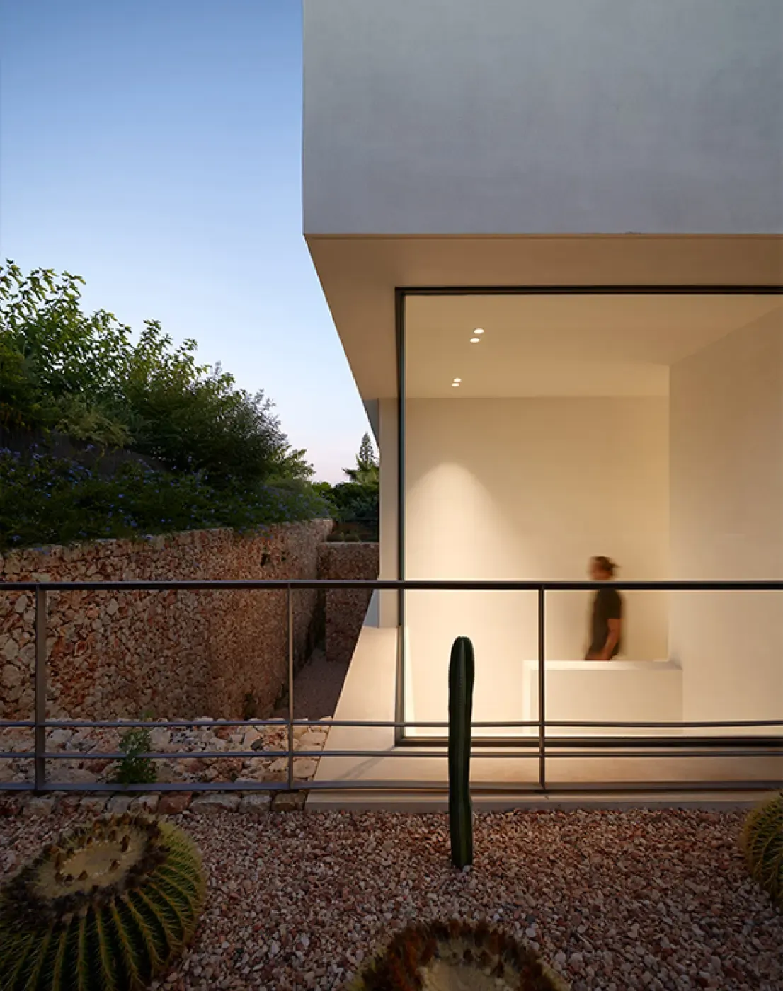 View of a minimalist concrete façade with floor-to-ceiling glass and a cactus garden at dusk in Menorca.