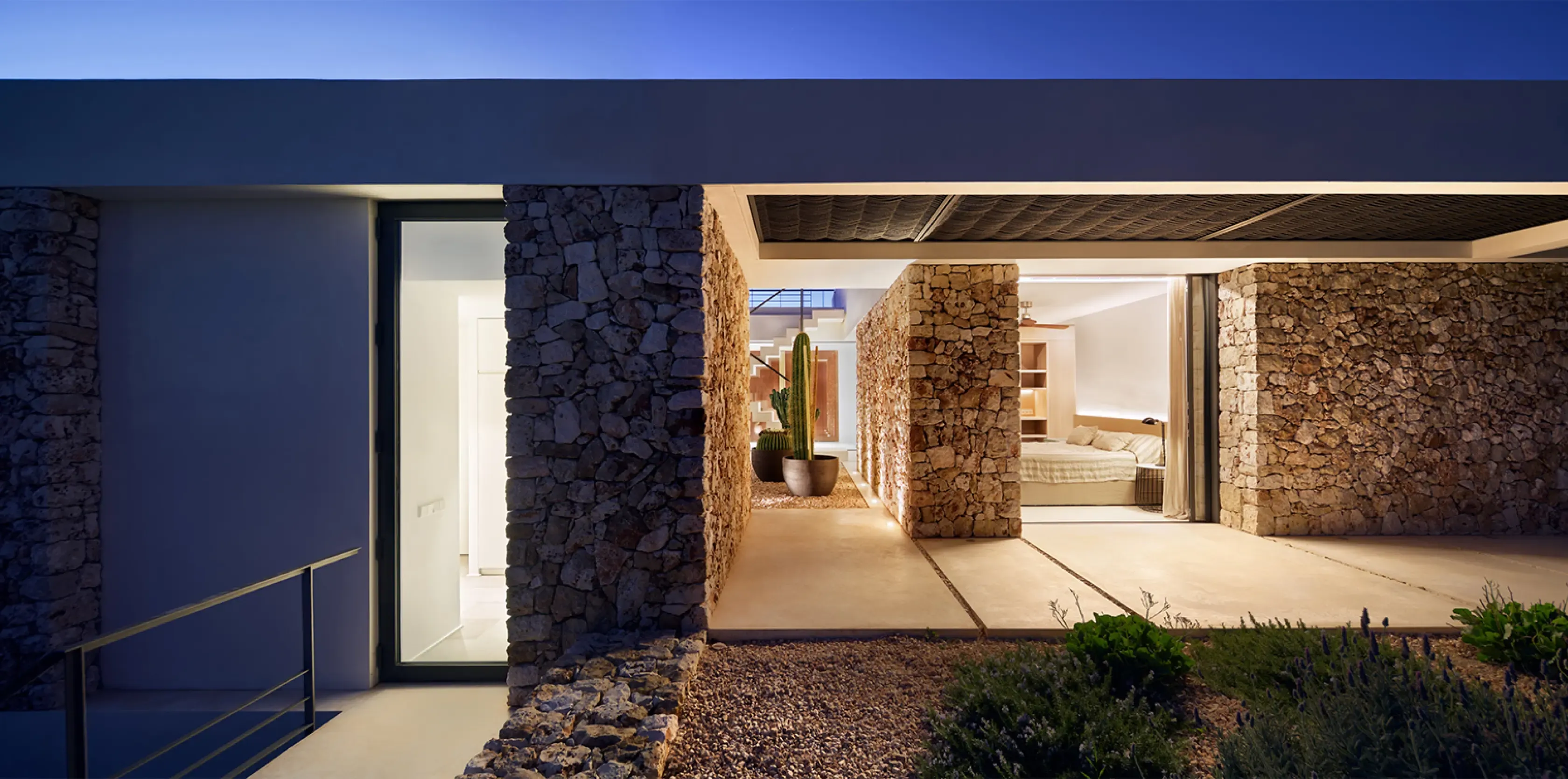 Night view of a stone-walled corridor leading to a bedroom, lit softly and flanked by desert plants.