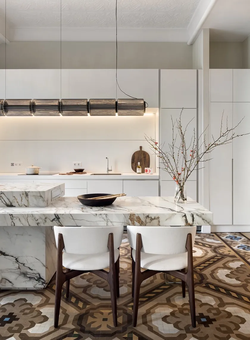 Detalle de la cocina de Casa Oller con sillas de madera y tapizado blanco, isla de acabado veteado e iluminación decorativa.
