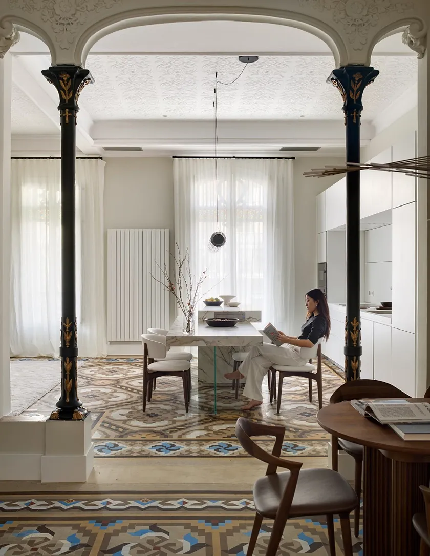 Vista de Casa Oller entre columnas modernistas ornamentadas, con mobiliario de cocina blanco, sillas tapizadas y suelo hidráulico.