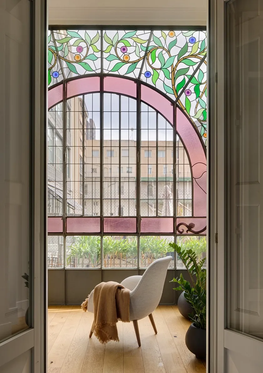 Galería modernista de Casa Oller con una butaca contemporánea tapizada en blanco, textiles cálidos y vidriera de colores.