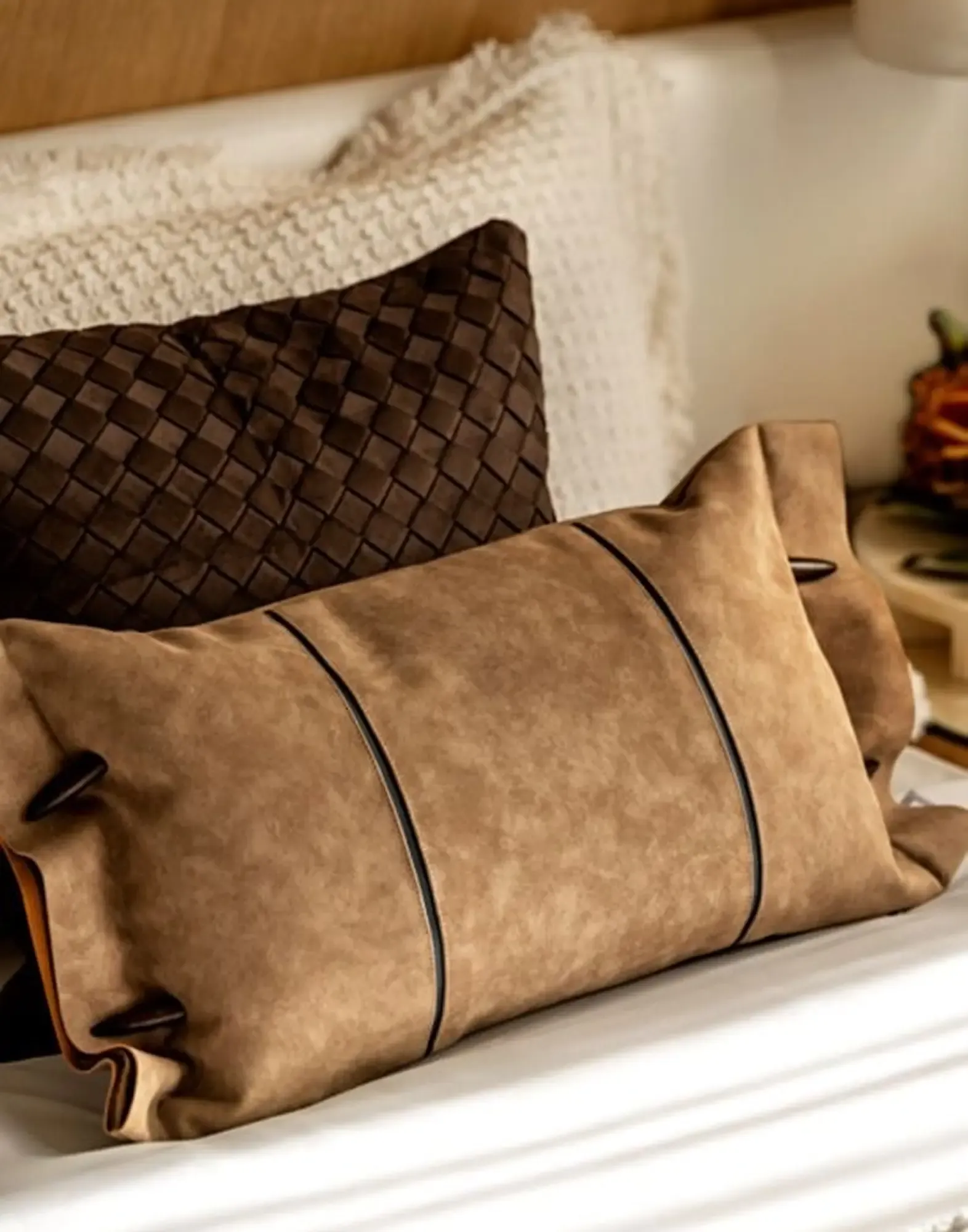 Decorative pillows in woven leather and tan suede textures on a neutral bed.