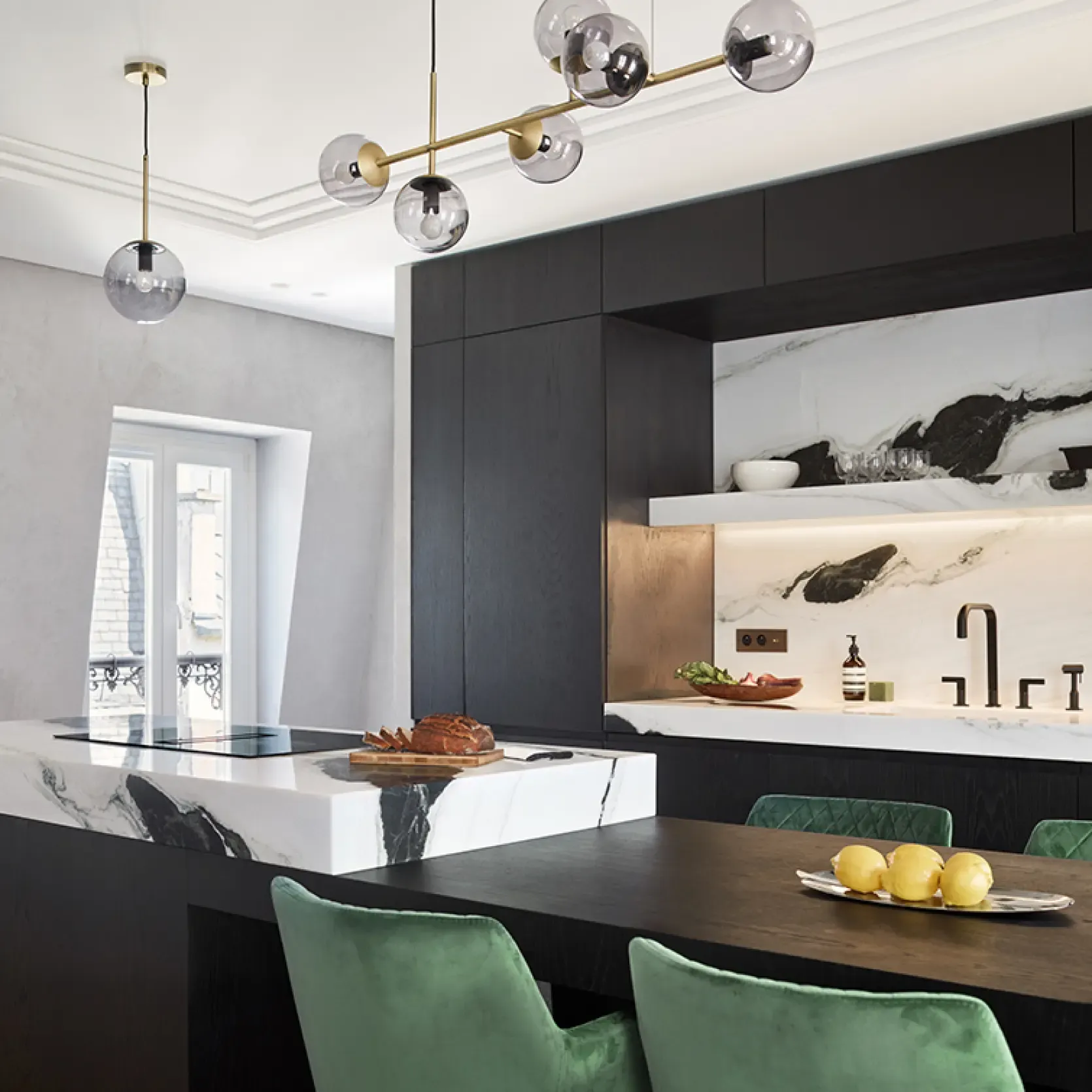 Kitchen interior with marble island, black cabinetry, and green velvet chairs highlighted by brass light fixtures and natural daylight.