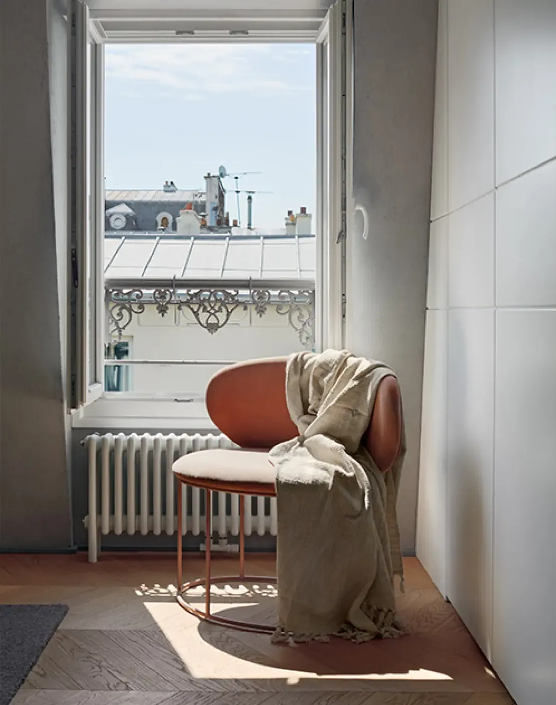 Cozy reading corner with terracotta armchair and linen throw beside a window overlooking Parisian rooftops and ornate balcony railings.
