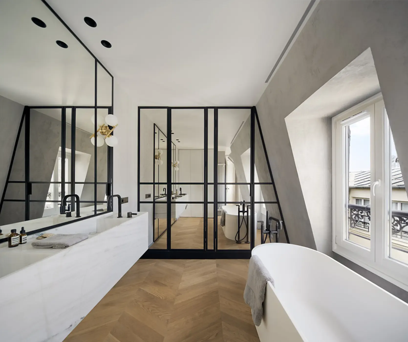 Bright bathroom featuring freestanding bathtub, marble double sink, glass partitions with black frames, and herringbone wooden floor.