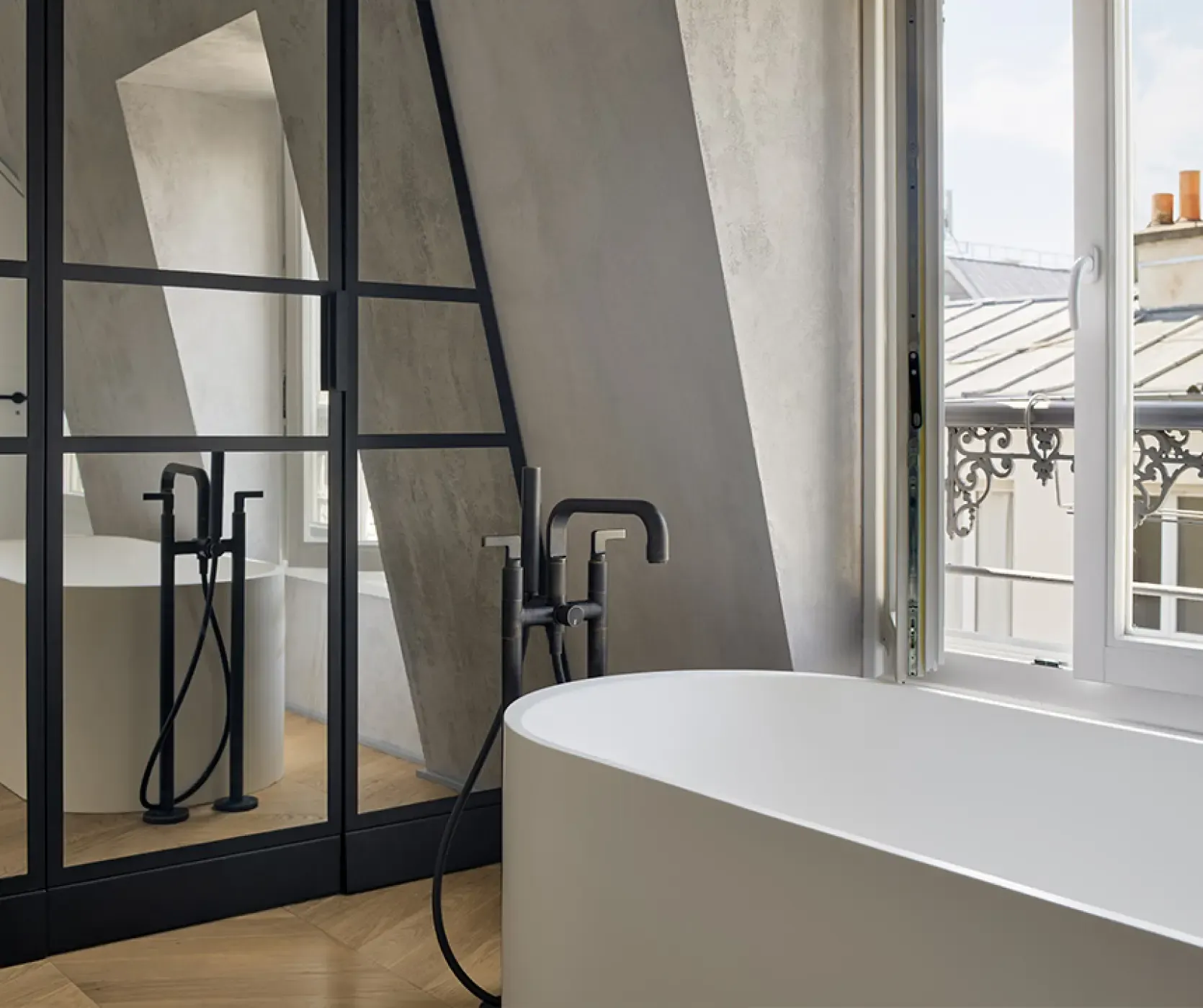 Detail of freestanding white bathtub with black fixtures placed beside a large window offering views of Paris rooftops and sky.
