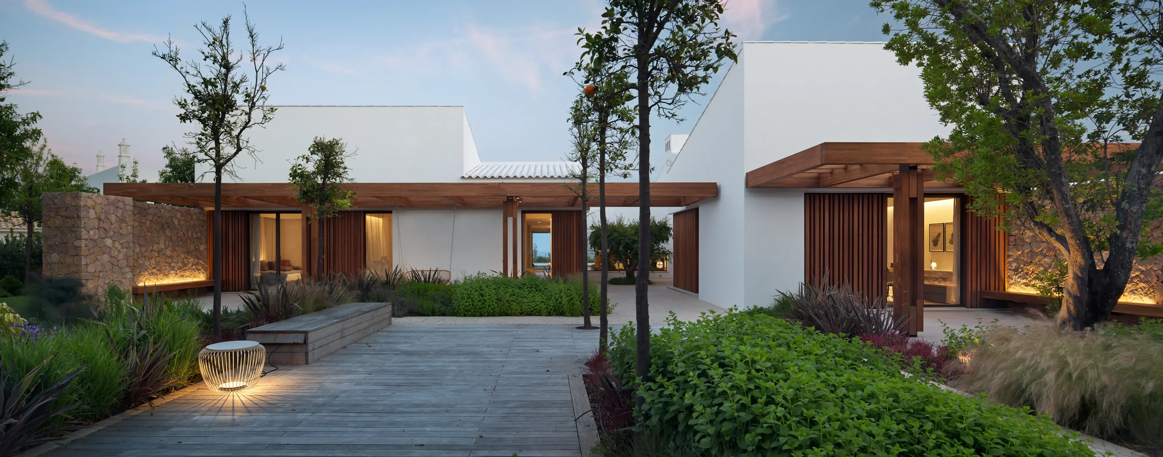 Exterior view at dusk with landscaped garden, illuminated stone walls, and wooden pergolas creating a warm inviting atmosphere.
