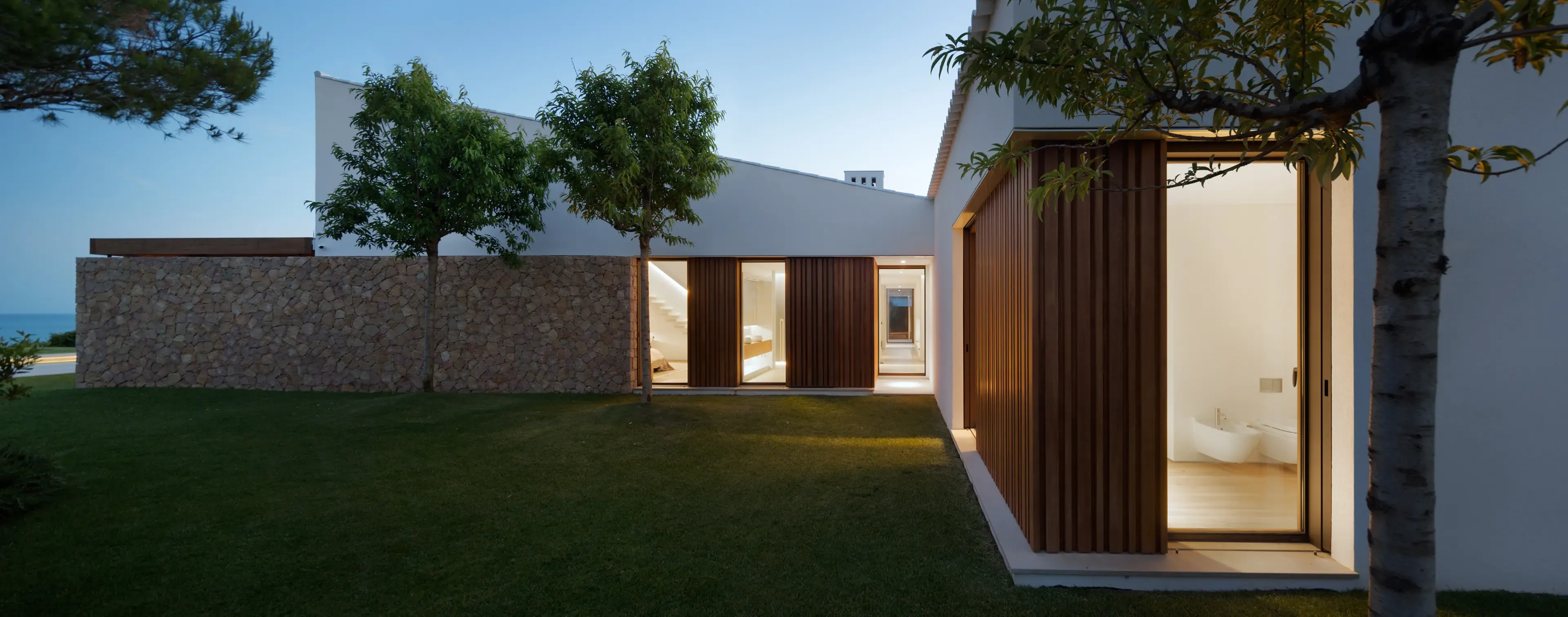 Evening exterior perspective with stone walls, green lawn, and large windows revealing softly lit interiors.