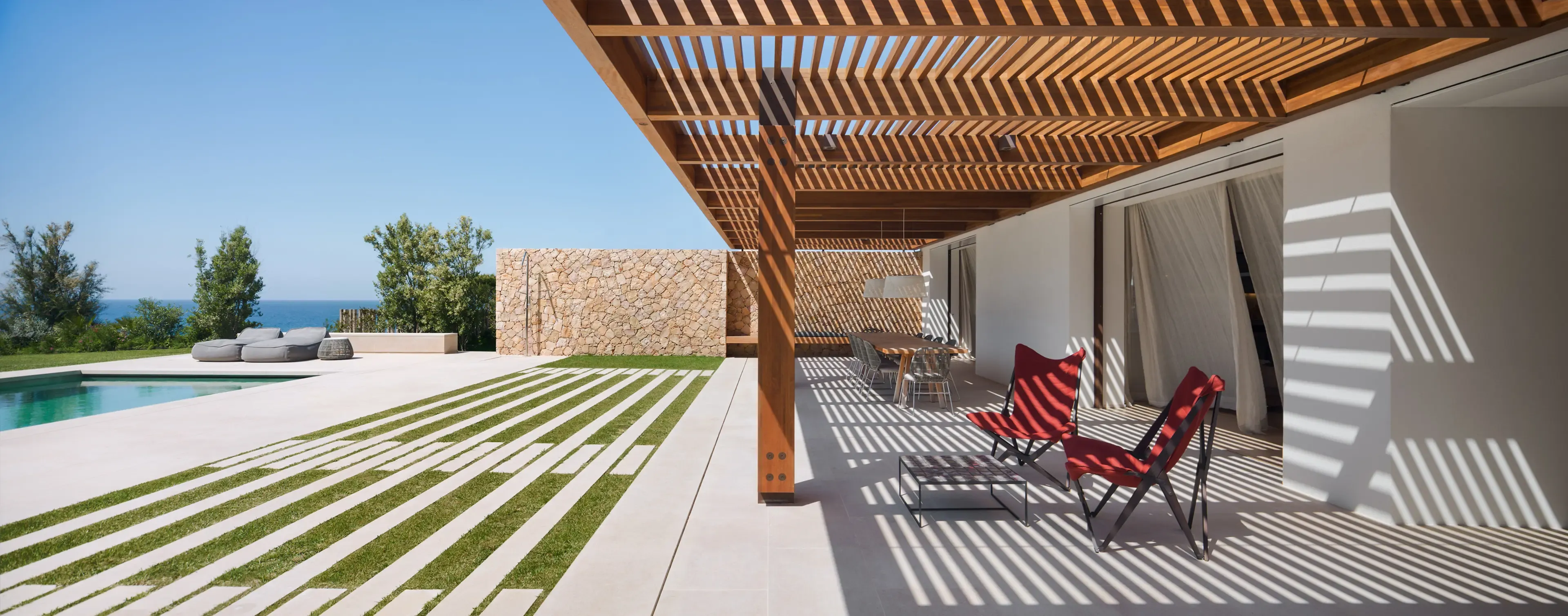 Shaded terrace with wooden pergola, red lounge chairs, and stone walls overlooking swimming pool and Mediterranean Sea.