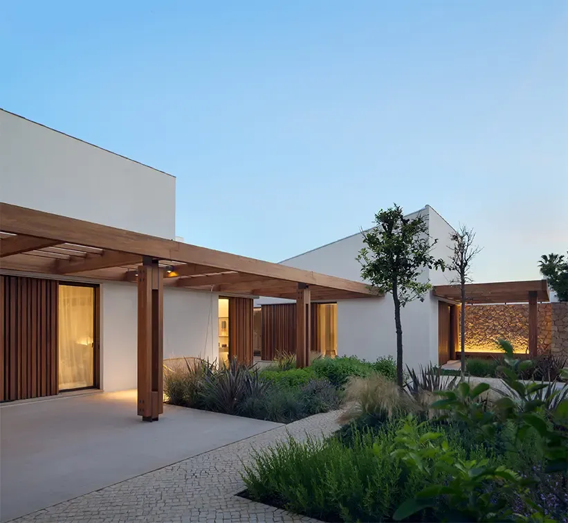 Contemporary villa façade at dusk with wooden pergolas, illuminated stone walls, and lush landscaped garden paths.