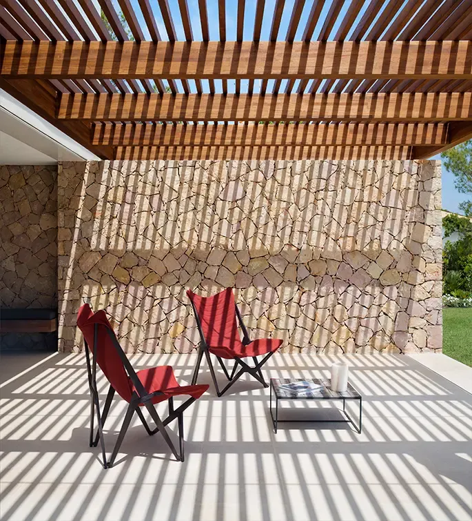 Outdoor seating corner with red chairs under wooden pergola casting shadows on rustic stone wall.