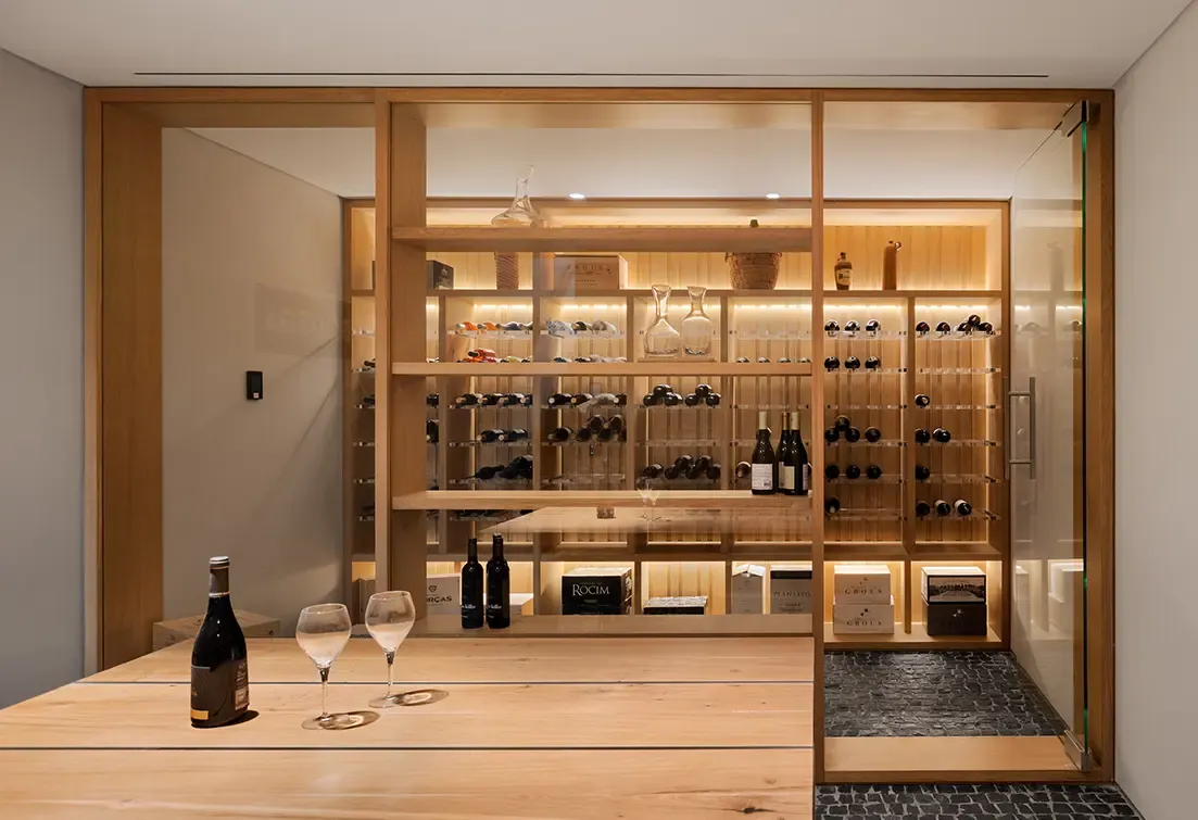 Illuminated wine cellar with glass enclosure, wooden shelving, and tasting table with bottles and glasses.