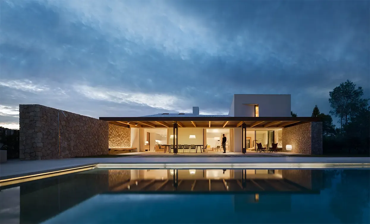 Evening exterior of luxury residence with illuminated interiors, expansive glass walls, stone facades, and swimming pool reflecting contemporary design.