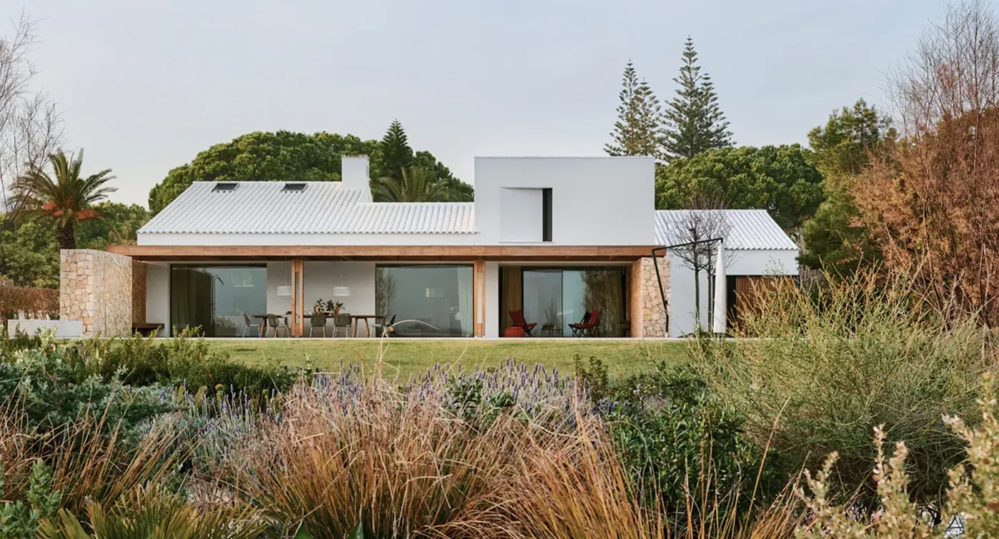 Modern villa exterior with white roof tiles, large glass windows, wooden pergolas, and landscaped Mediterranean garden.