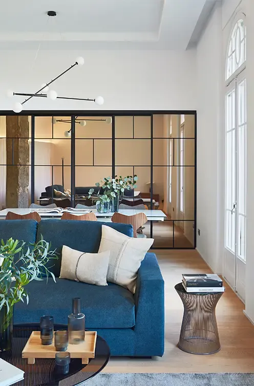 Stylish lounge with blue sofa, designer chandelier, glass partition, and dining area, showcasing elegant open-plan apartment architecture.