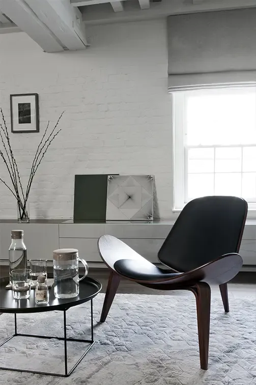 Stylish corner view of lounge chair and coffee table with glassware, set against a white brick wall and large window in a modern apartment.