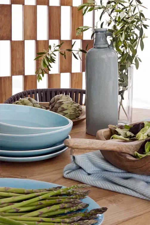 Detail of outdoor dining setup with blue tableware, artichokes, and fresh salad against wooden lattice backdrop.