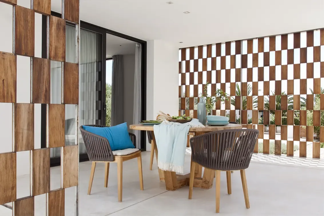 Covered outdoor dining area with wooden table, woven chairs, and lattice wall blending indoor-outdoor living.