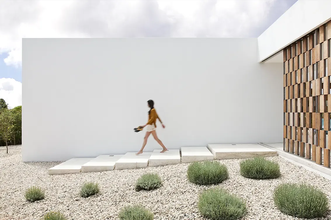 Side view of modern white facade with pathway steps and woman walking across gravel garden.
