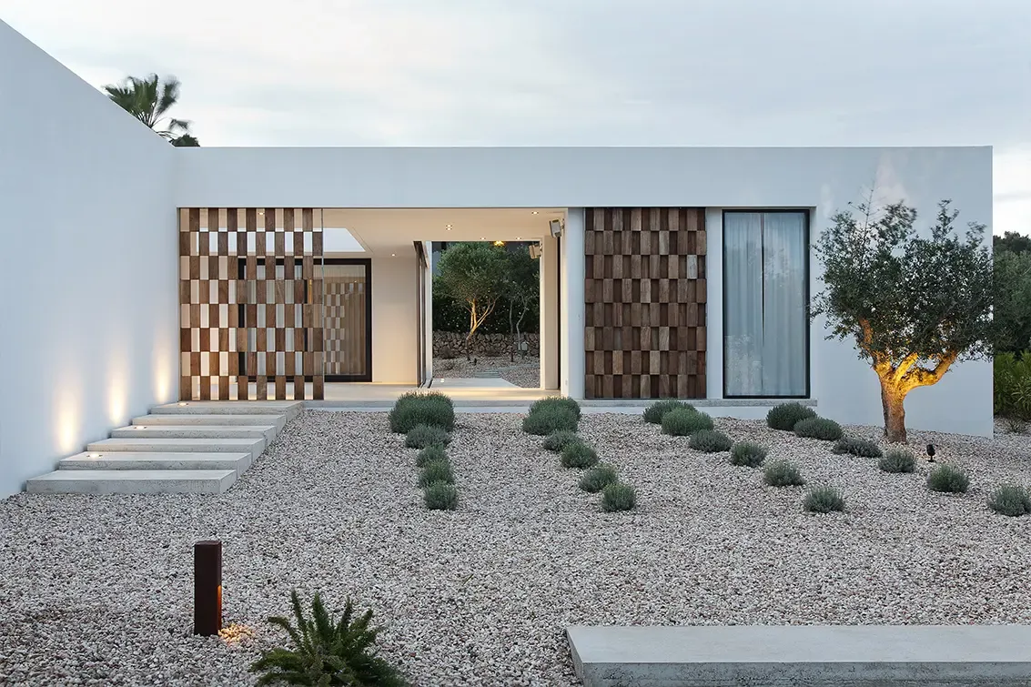 Evening view of house entrance with illuminated lattice panels and olive tree creating warm atmosphere.