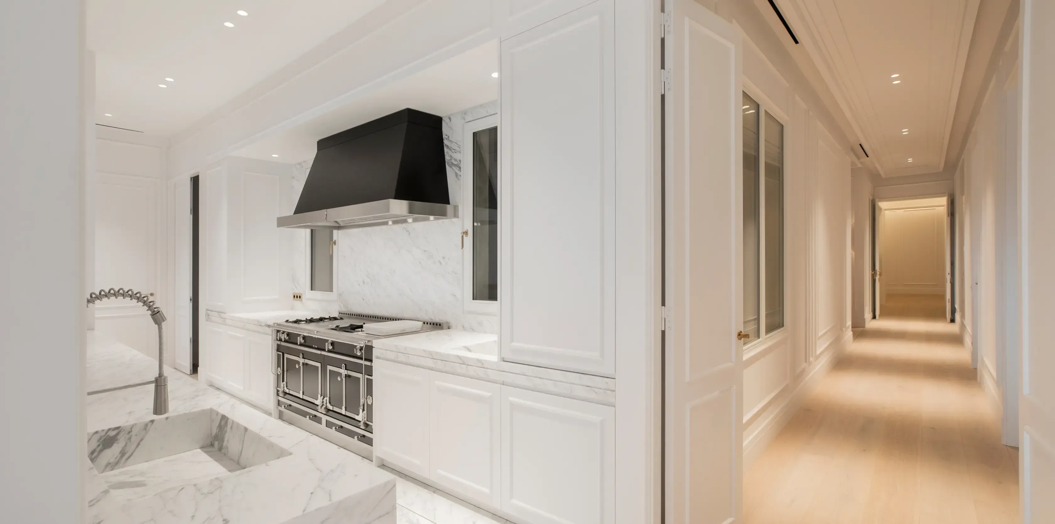 Bright kitchen with marble countertops, a large black hood, and white cabinetry connecting to a warm wood-floor hallway.
