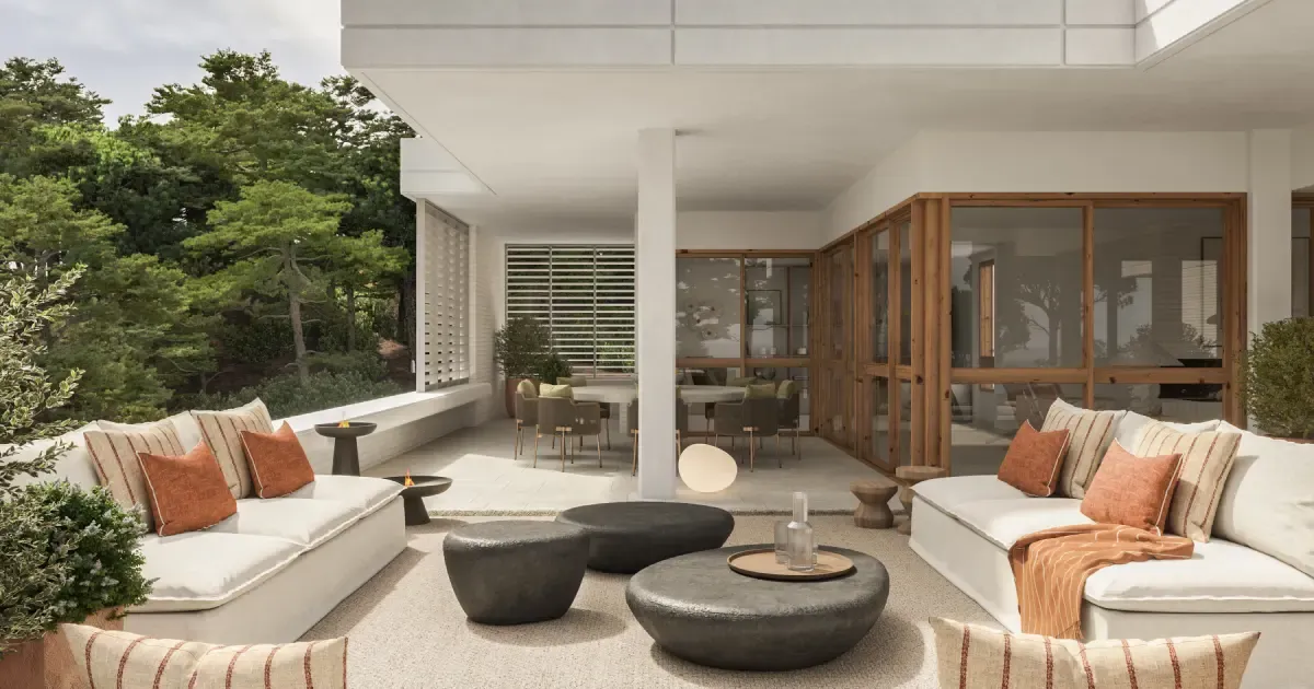 Outdoor lounge on a luxury terrace with natural tones, looking toward a modern villa facade surrounded by forest.